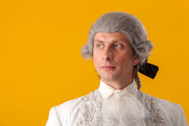 Retro baroque man with white wig holding a walking stick sitting on antique couch. Studio shot against blue.