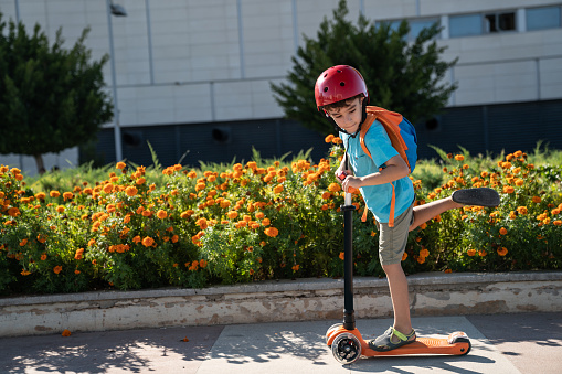 Little boy wearing a striped t-shirt, blue jeans and a red helmet using push scooter in outdoor.The background is blurred.