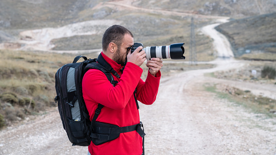 Adult photographer man wearing a red coat photographing in rural region. Dirt country road is seen on the background. Shot in outdoor with a full frame mirrorless camera.