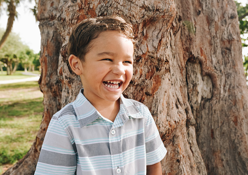 Cute little boy, 5 years old, mixed race, outdoors in front of a tree in a candid portrait, full of joy and childhood smiles