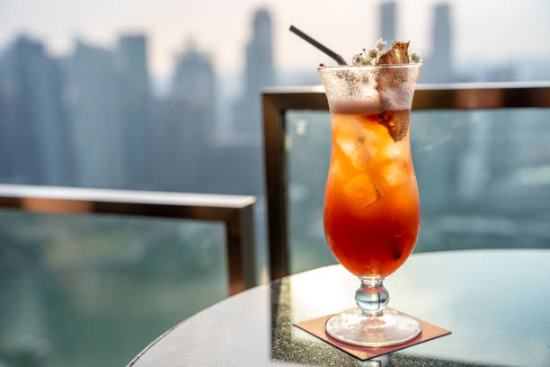 Orange and yellow cocktail with floral garnish in a tall curvy glass with condesnation on a table in a roof top patio with bokeh cityscape in the background