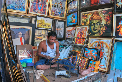 Front view of a photo framing shop in Janbazar, a neighbourhood of central Kolkata. A shop owner / skilled person in seen working in one of the shops, in midst of several photo frames hanging in wall. Some of the working tools can also be seen.\n\nJanbazar is famous for photo framing shops and residence of Rani Rasmoni, who was a pillar of strength in the male dominated society of mid-nineteenth century Bengal in India.