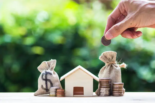 Photo of Saving money, home loan, mortgage, a property investment for future concept : A man hand putting money coin over small residence house and money bag.