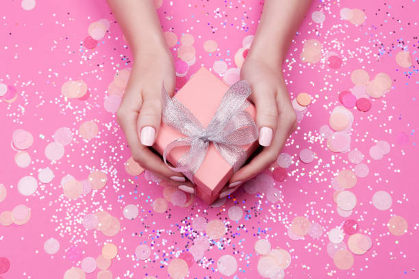 istock Woman hands holding present box with silver bow on pastel pink background with glitter and confetti. Flat lay style. 1057833238