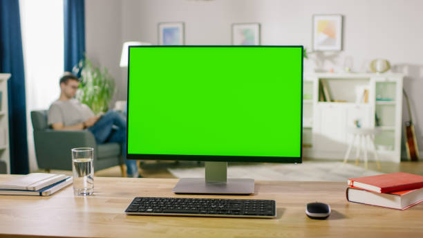 istock Modern Personal Computer with Green Mock-up Screen Display Standing on the Desk. In the Cozy and Light Living Room Man Relaxes on a Chair Reading a Book. 1057833314
