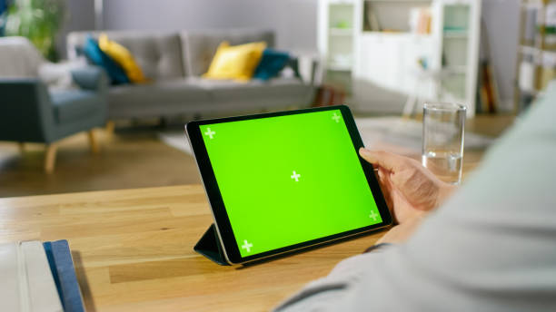 istock Man Holding and Watching Green Mock-up Screen Digital Tablet Computer While Sitting at the Desk. Man Watching Videos or Browsing Through the Internet. In the Background Cozy Living Room. 1057833356