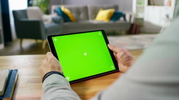 istock Man Holding and Watching Green Mock-up Screen Digital Tablet Computer While Sitting at the Desk. Man Watching Videos or Browsing Through the Internet. In the Background Cozy Living Room. 1057833358