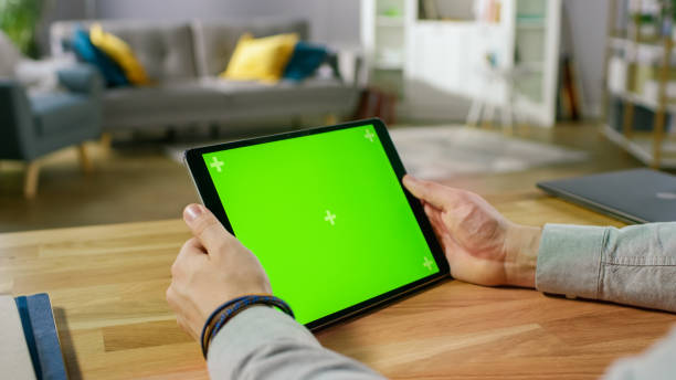 istock Man Holding and Watching Green Mock-up Screen Digital Tablet Computer While Sitting at the Desk. Man Watching Videos or Browsing Through the Internet. In the Background Cozy Living Room. 1057833360