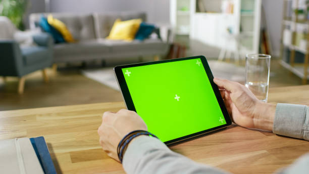 istock Man Holding Green Mock-up Screen Digital Tablet Computer While Sitting at the Desk. Man Buying stuff or Browsing Through the Internet. In the Background Cozy Living Room. 1057833366