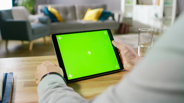 istock Man Holding Green Mock-up Screen Digital Tablet Computer While Sitting at the Desk. Man Buying stuff or Browsing Through the Internet. In the Background Cozy Living Room. 1057833368