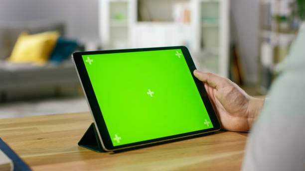 istock Man Watching Green Mock-up Screen Digital Tablet Computer While Sitting at the Desk. Man Watching Videos or Browsing Through the Internet. In the Background Cozy Living Room. 1057833376