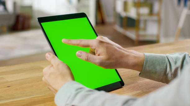 istock Man Using Hand Gestures on Green Mock-up Screen Digital Tablet Computer in Portrait Mode while Sitting at His Desk. In the Background Cozy Living Room. 1057833394