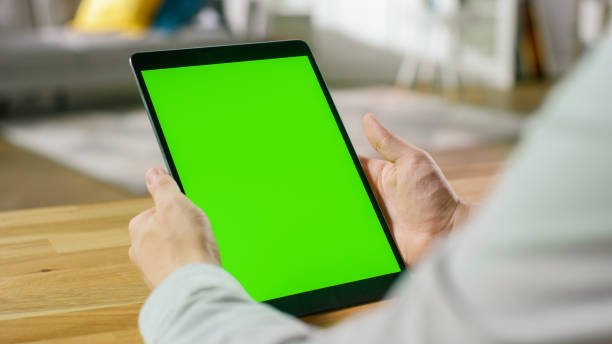 istock Man Using Green Mock-up Screen Digital Tablet Computer in Portrait Mode while Sitting at His Desk. In the Background Cozy Living Room. 1057833396