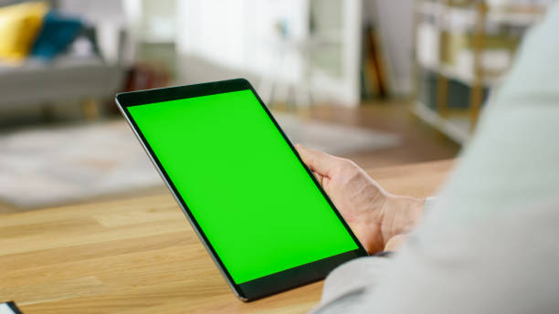 istock Man Using Green Mock-up Screen Digital Tablet Computer in Portrait Mode while Sitting at His Desk. In the Background Cozy Living Room. 1057833402