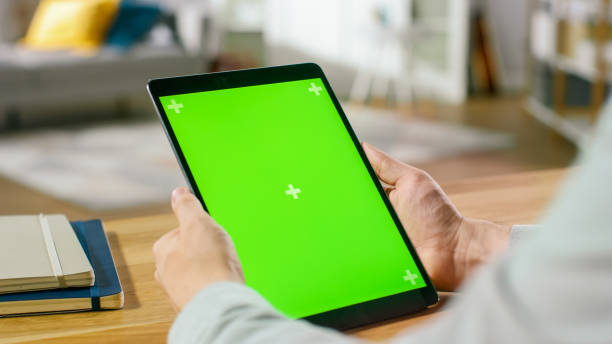 istock Man Using Green Mock-up Screen Digital Tablet Computer in Portrait Mode while Sitting at His Desk. In the Background Cozy Living Room. 1057833412