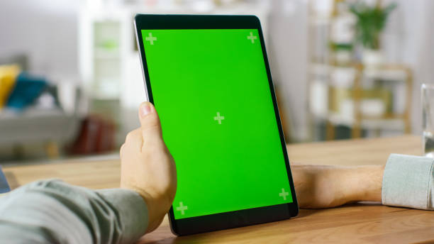 istock Close-up of Man Using Hand Gestures on Green Mock-up Screen Digital Tablet Computer in Portrait Mode while Sitting at His Desk. In the Background Cozy Living Room. 1057833424