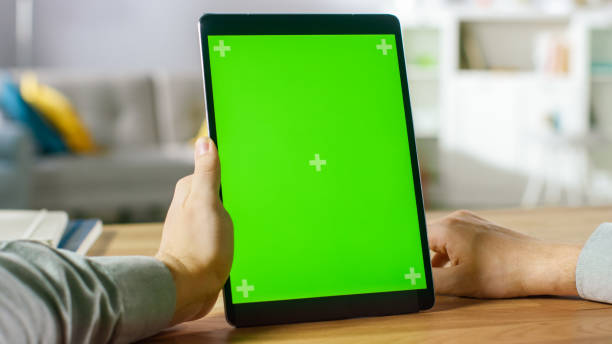 istock Close-up of Man Using Hand Gestures on Green Mock-up Screen Digital Tablet Computer in Portrait Mode while Sitting at His Desk. In the Background Cozy Living Room. 1057833426