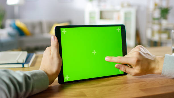 istock Close-up of Man Using Hand Gestures on Green Mock-up Screen Digital Tablet Computer in Landscape Mode while Sitting at His Desk. In the Background Cozy Living Room. 1057833436