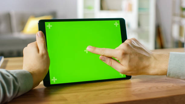 istock Close-up of Man Using Hand Gestures on Green Mock-up Screen Digital Tablet Computer in Landscape Mode while Sitting at His Desk. In the Background Cozy Living Room. 1057833444