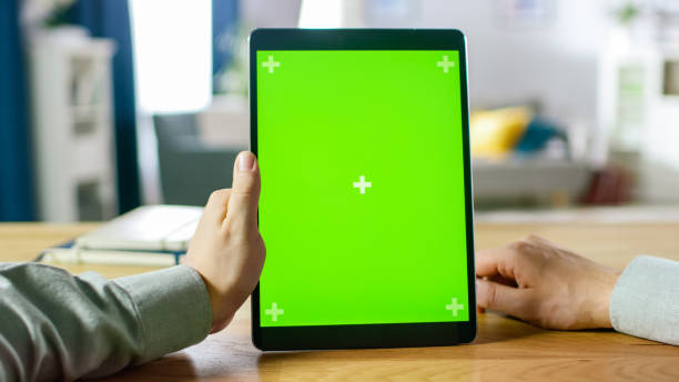 istock Close-up of Man Using Hand Gestures on Green Mock-up Screen Digital Tablet Computer in Portrait Mode while Sitting at His Desk. In the Background Cozy Living Room. 1057833446