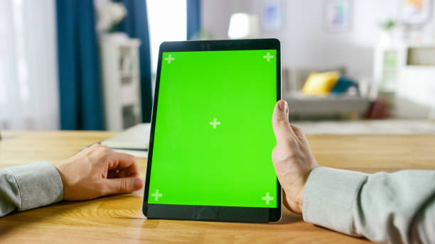istock Close-up of Man Using Green Mock-up Screen Digital Tablet Computer in Portrait Mode while Sitting at His Desk. In the Background Cozy Living Room. 1057833458