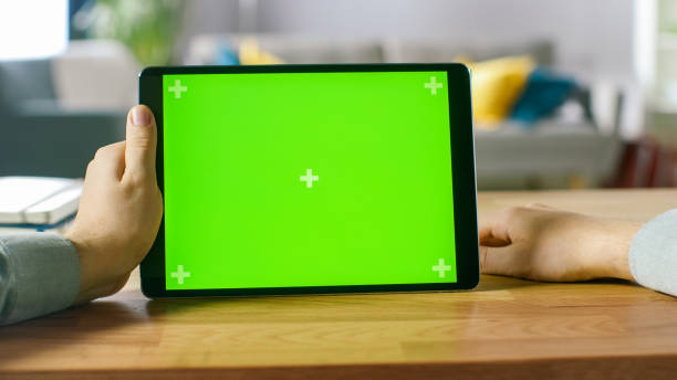 istock Man Holding and Watching Green Mock-up Screen Digital Tablet Computer While Sitting at the Desk. Man Watching Videos in Landscape Mode. 1057833460