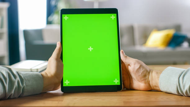 istock Close-up of Man Using Green Mock-up Screen Digital Tablet Computer in Portrait Mode while Sitting at His Desk. In the Background Cozy Living Room. 1057833462