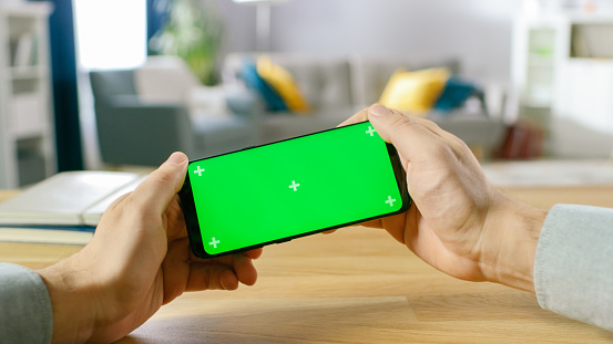 First Person Close-up of the Man Holding Green Screen Smartphone in Landscape Mode and Playing in a Racing Video Game by Tilting Mobile Phone. Background Cozy Living Room.