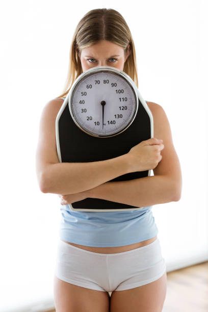 istock Frustrated young woman looking at camera with weigh scale over white background. 1057844732