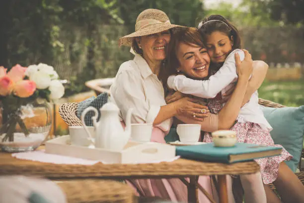 Three generation of women are hugging on a garden sofa. They are celebrating Mother's day by having an outdoor tea party.
