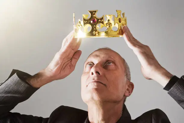 A mature man looks upwards as he crowns himself with a shiny golden coronet - obviously with a high opinion of himself.