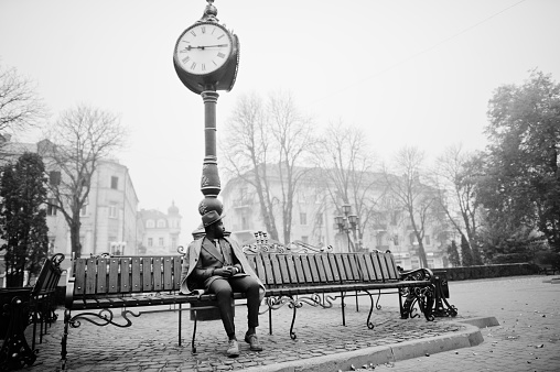 Stylish African American man model in gray coat, jacket tie and red hat posed at bench against large clock. Black and white photo.