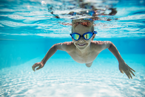 Portrait of smiling little boy swimming underwater in the pool towards the camera. Sunny summer day.\nNikon D850