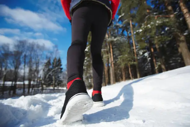 Legs of sportswoman in black leggins and cross-shoes jogging on snowpath in winter forest