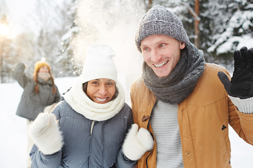Cheerful excited young multi-ethnic couple in warm scarves and hats protecting from snow thrown by friends while walking in forest