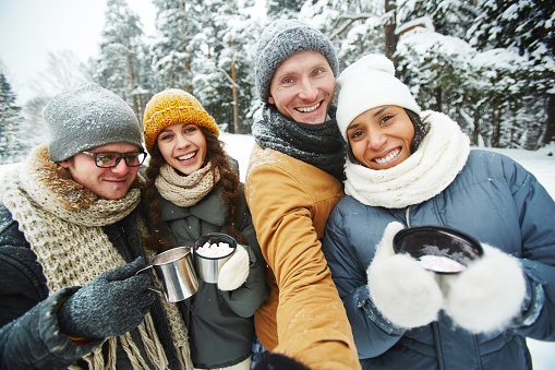 Positive excited young multi-ethnic friends in warm clothing holding thermos mugs and taking selfie together in winter forest, they hiking together