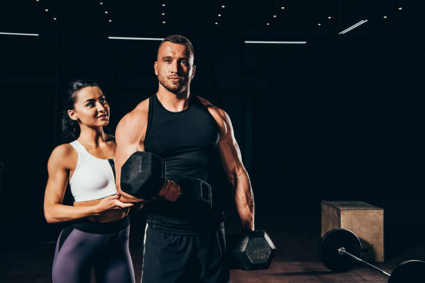 istock athletic bodybuilder exercising with barbell and looking at camera while woman standing behind in dark gym 1057945818