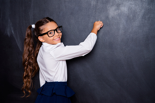 Portrait of nice genius cheerful adorable lovely small little girl with curly pigtails in white formal blouse shirt, blue skirt, writing on blackboard. Isolated over black background