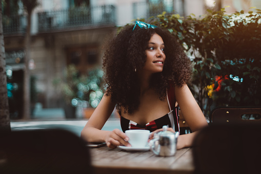 A cheerful cute curly girl is looking aside while sitting alone in a cafe outdoors with a cup of hot tea; young African-American female in street bar drinking cappuccino while waiting for her friend