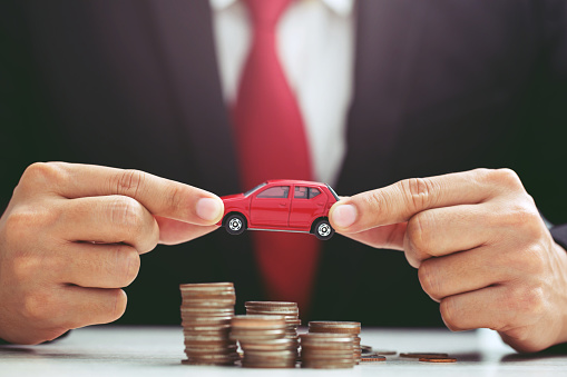 Business man in suit and red necktie holding model of toy car on over a lot of stacked coins - insurance, loan and buying car concept
