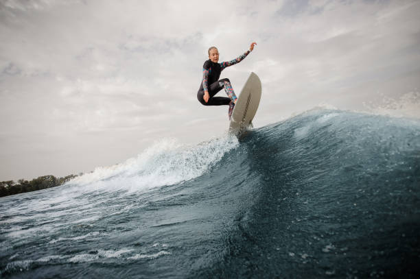 Young slim and active woman in swimsuit jumping up blue wave against the grey sky Young slim and active woman in swimsuit jumping up on the wakeboard against the grey sky wave jumping stock pictures, royalty-free photos & images