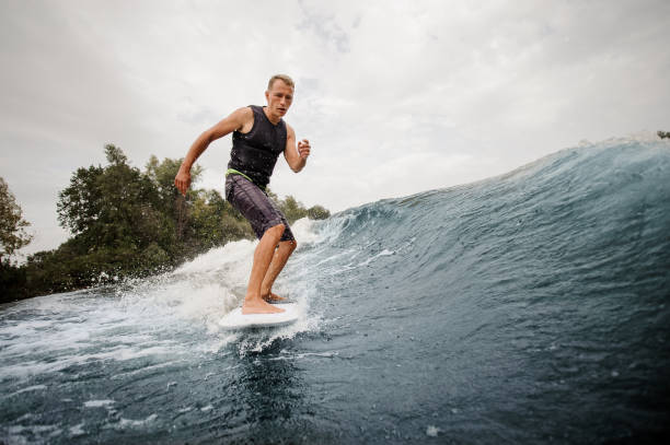 istock Active young man wakesurfing on the board down the blue high wave against grey sky 1058062922