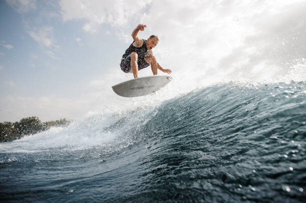 giovane uomo attivo che salta sul wakeboard bianco lungo l'acqua blu contro il cielo grigio - wakeboarding surfing men vacations foto e immagini stock