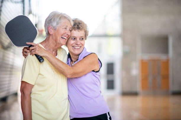 Senior friends spending time together Two (2) senior friends hug each other while sharing a laugh. They are having a relaxing and joyful day at the gym playing pickleball. pickleball stock pictures, royalty-free photos & images