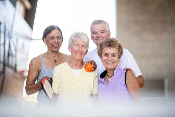 Four (4) senior friends face forward smiling. One older woman rests her hand around her friend. They are all happy after exercising.