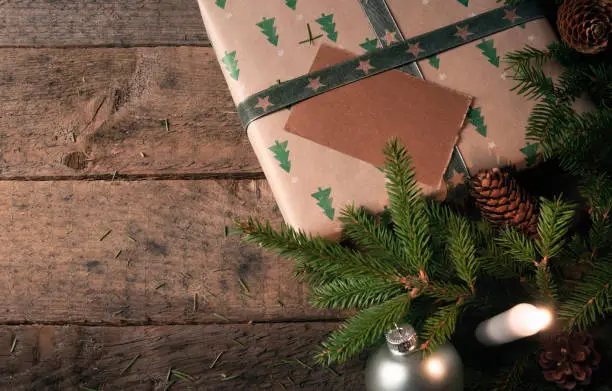 Green fir twigs, pine cones, and Christmas balls, near a gift wrapped in classic brown paper with trees, and a lit candle, on a vintage wooden table.