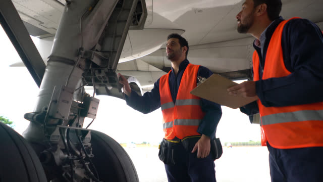 Team of airplane mechanics checking the landing gear of an airplane one points at parts and the other one takes notes