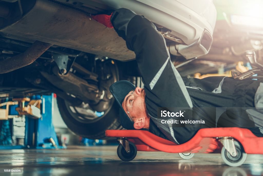 Car Mechanic Under the Car Caucasian Car Mechanic in His 30s Under the Car on the Mechanics Creeper Trying To Fix Modern Vehicle Exhaust System. Car Stock Photo