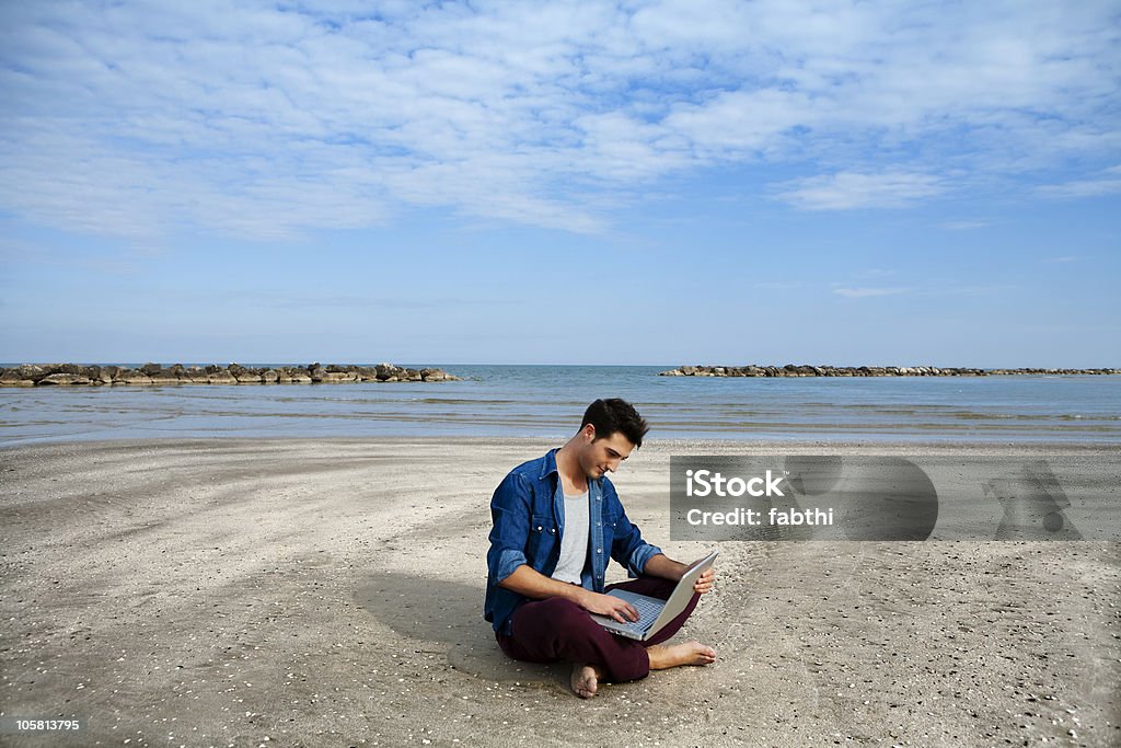 Jeune homme assis sur la plage avec ordinateur portable - Photo de 20-24 ans libre de droits