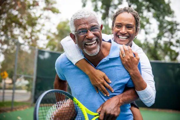 A senior black couple piggyback together on the tennis court.